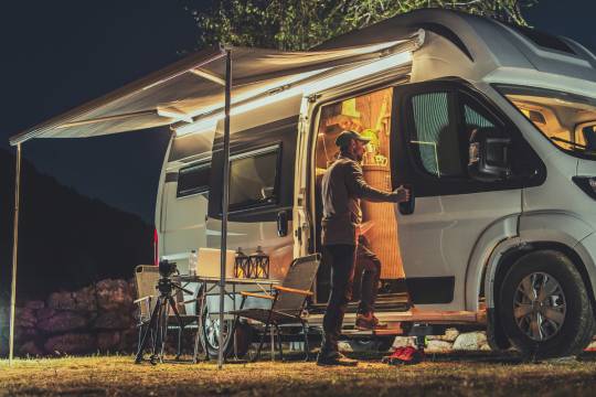 Homme entrant dans un van aménagé illuminé la nuit, camping avec auvent déployé et matériel photo installé en extérieur