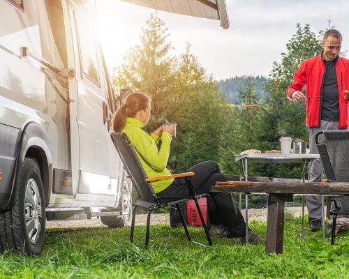Couple profitant du camping en van, avec un homme grillant des saucisses et une femme savourant une boisson chaude.