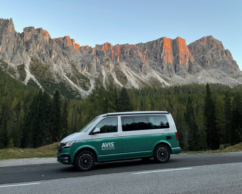 Van aménagé AVIS explore circulant sur une route de montagne avec une vue des cimes au coucher du soleil