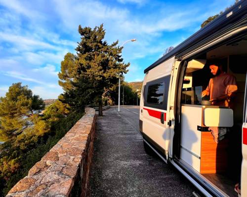 Homme cuisinant à l'intérieur d'un van aménagé AVIS explore, garé le long d'une route panoramique bordée d'arbres et surplombant la mer.