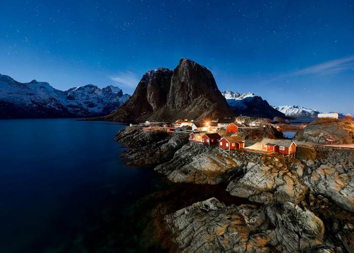 Village de pêcheurs de Hamnøy aux îles Lofoten, Norvège, avec ses maisons rouges traditionnelles nichées entre montagnes enneigées et fjords, sous un soleil scintillant et un ciel saisissant
